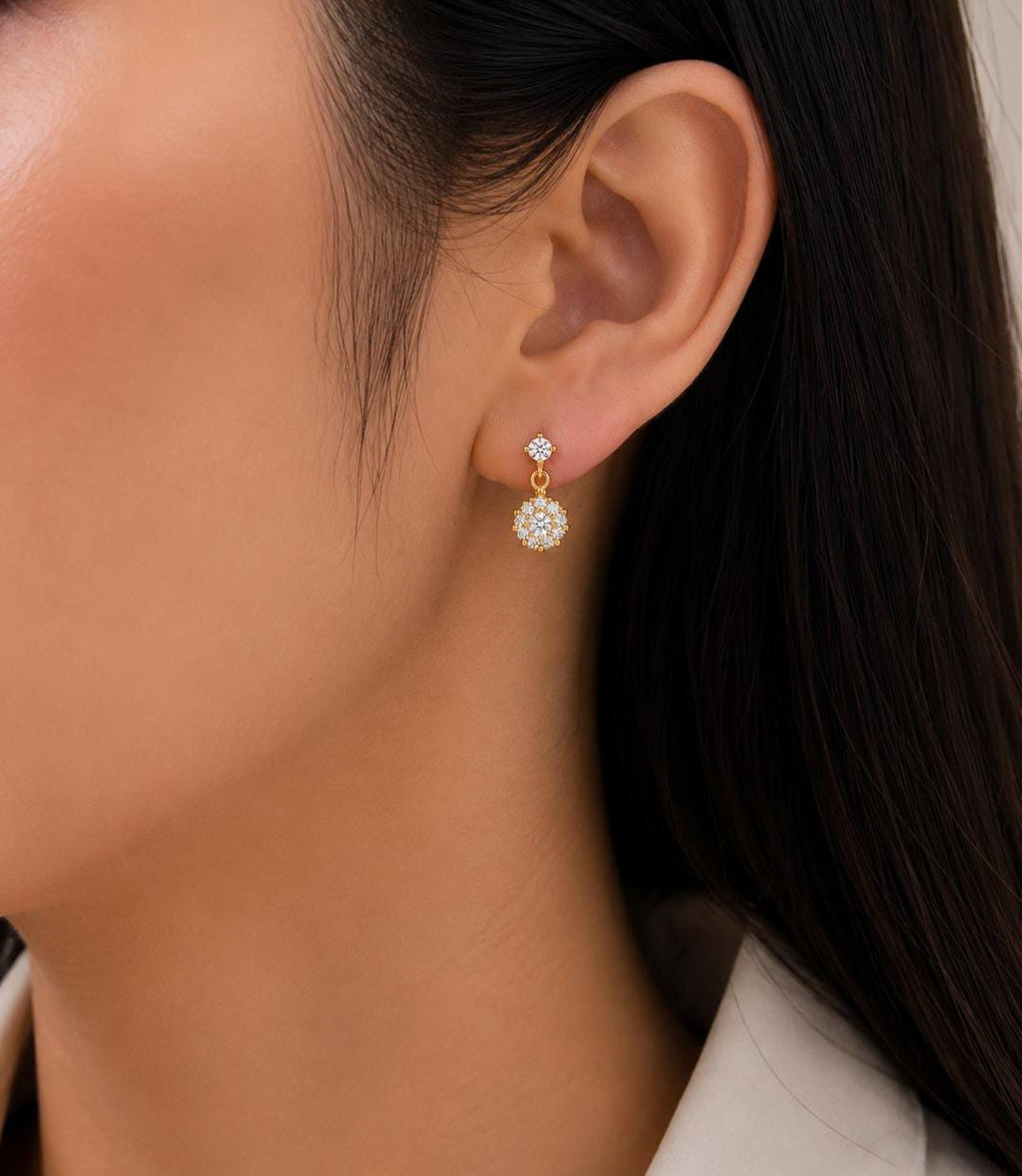 A close-up of a model's ear wearing a delicate gold dangle earring with a small clear gemstone and a larger round pavé-set clear stone drop.