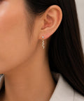 A close-up of a model's ear wearing a delicate silver dangle earring with a small clear stone and a wavy zigzag drop.