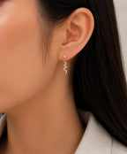A close-up of a model's ear wearing a delicate silver dangle earring with a small clear stone and a wavy zigzag drop.