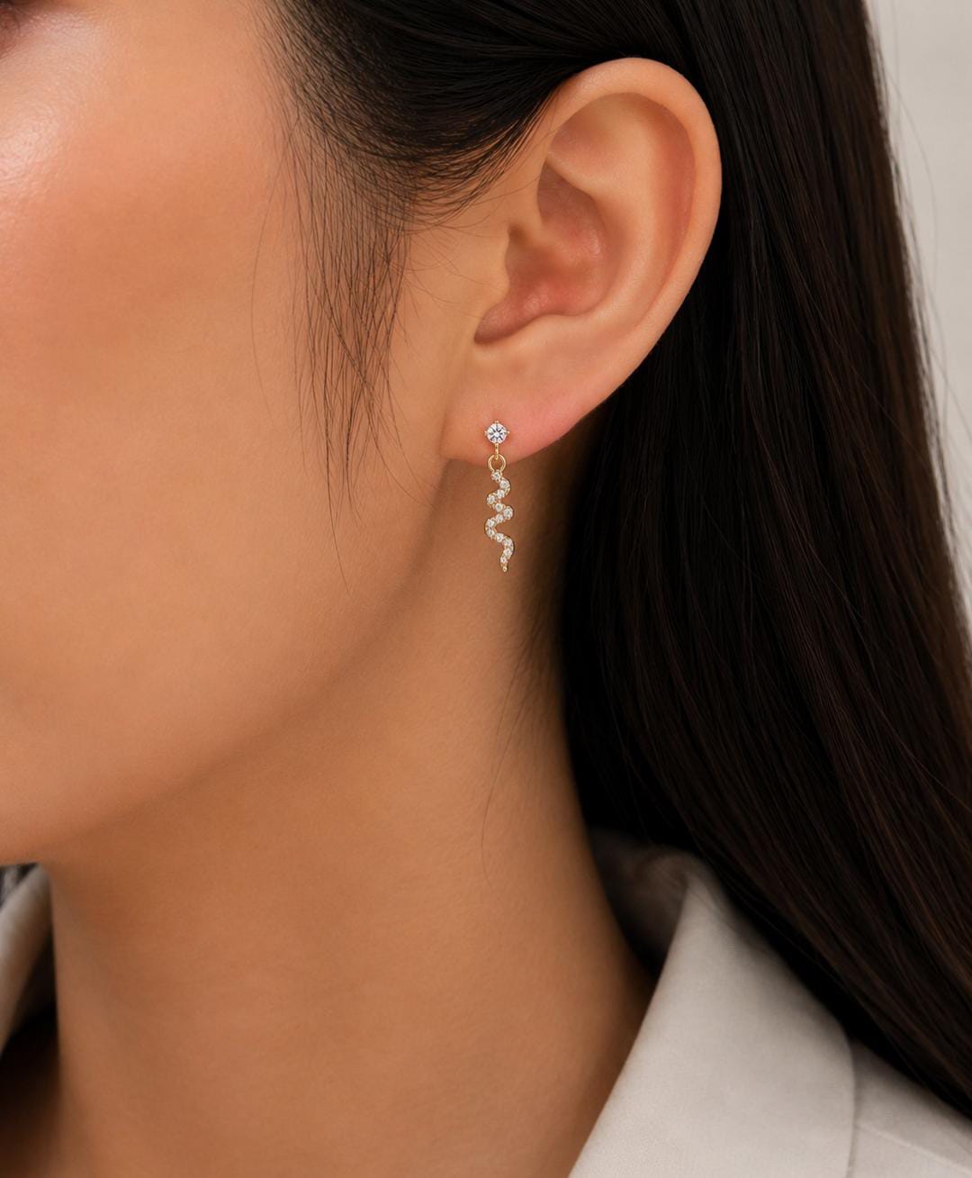 A close-up of a model's ear wearing a delicate silver dangle earring with a small clear stone and a wavy zigzag drop.